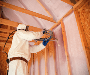 Worker in protective gear applying spray foam insulation to wooden framing.