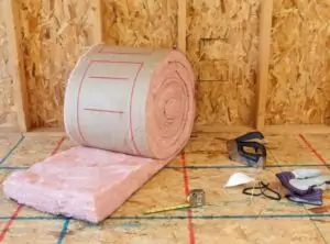 Roll of fiberglass batt insulation alongside the tools necessary for installing it, including tape measure, staple gun, and personal protective equipment.