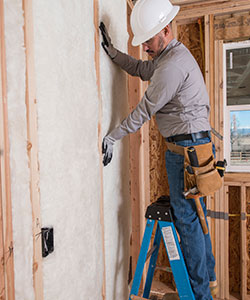 Worker installing white fiberglass batt insulation on an interior wall.