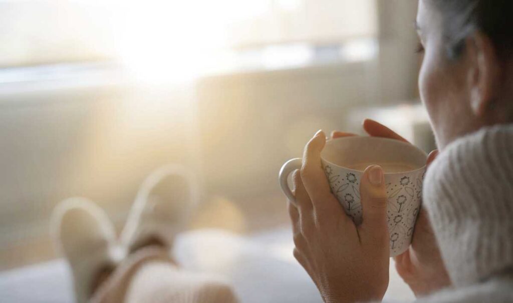Cozy woman drinking from a warm mug while enjoying a winter sunrise from indoors.